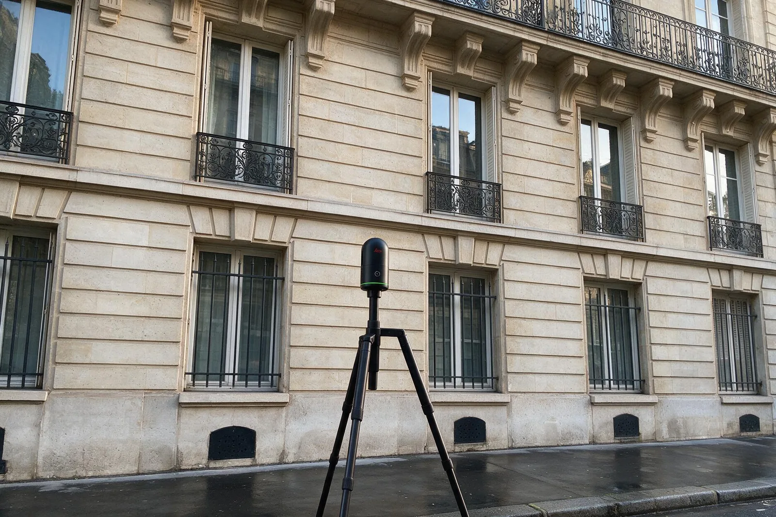 Scanner laser 3D statique sur trepied devant une facade en pierre de taille : garde-corps en ferronnerie, encadrements moulurés, corniche a modillons.